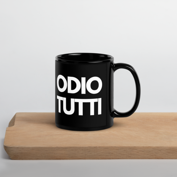 Un primo piano di una tazza in ceramica nera con la scritta ironica "Odio Tutti" stampata in caratteri bold. La tazza ha una finitura lucida ed è posizionata su un tavolo accanto a una moka e una tazza di caffè fumante. L'illuminazione calda crea un’atmosfera accogliente, perfetta per la colazione. Sullo sfondo, un libro aperto e un paio di occhiali suggeriscono un momento di relax o una pausa caffè.